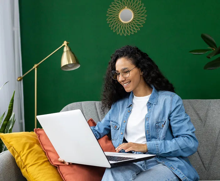 A woman with long curly hair and glasses sits on a grey couch, smiling while using a laptop. She wears a denim jacket and white t-shirt. There are colorful cushions and a green wall with gold decor behind her.