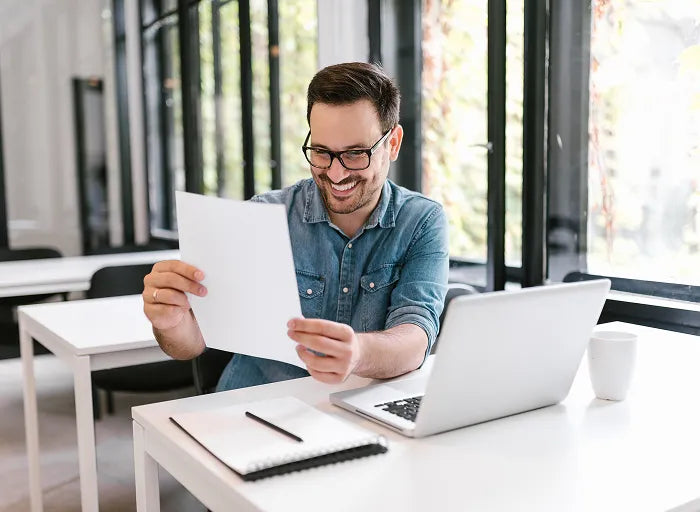 A man wearing glasses and a blue shirt sits at a desk with a laptop, notebook, and coffee cup, smiling while looking at a sheet of paper in his hands in a bright office space.