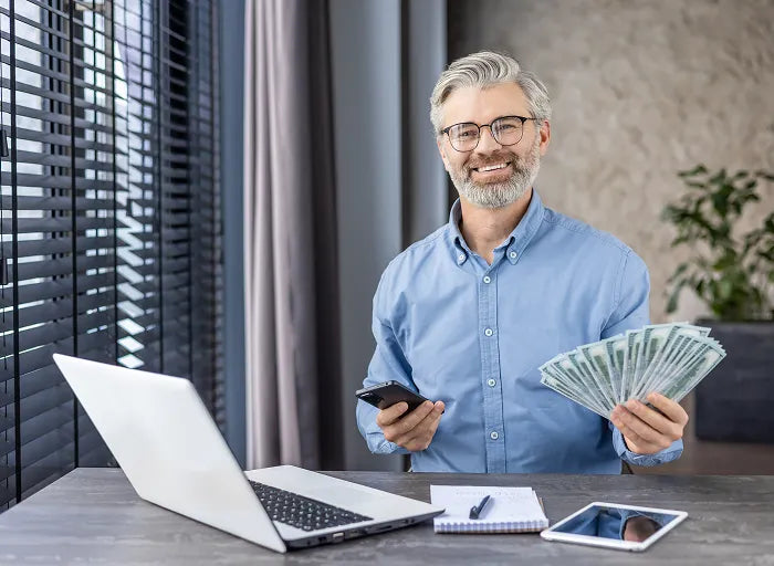 A smiling man with glasses sits at a desk holding a phone and a fan of cash. A laptop, notepad, pen, and tablet are on the desk. Light from window blinds shines on him.