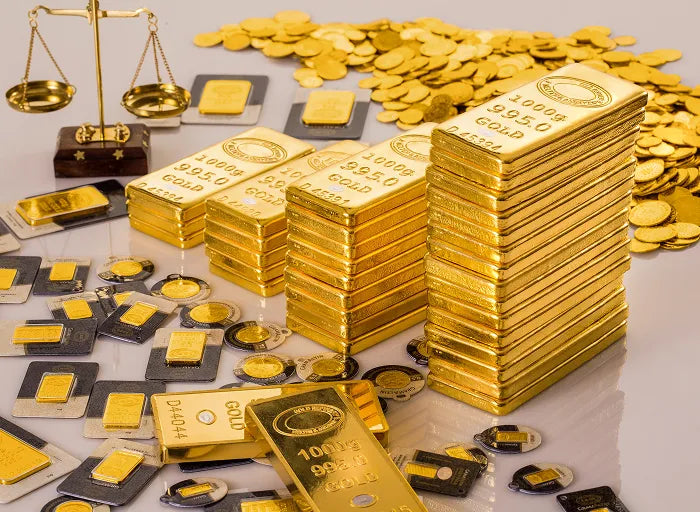 Stacks of gold bars, gold coins, and gold wafers are displayed on a reflective surface. A small scale balances gold coins in the background, suggesting themes of wealth and value.